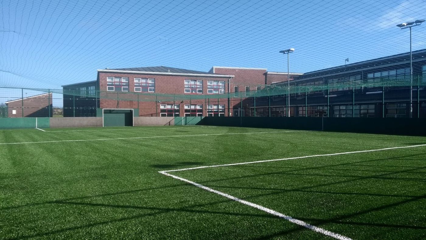 An artificial turf soccer field with white boundary lines, enclosed by tall nets, in front of a modern red-brick building.