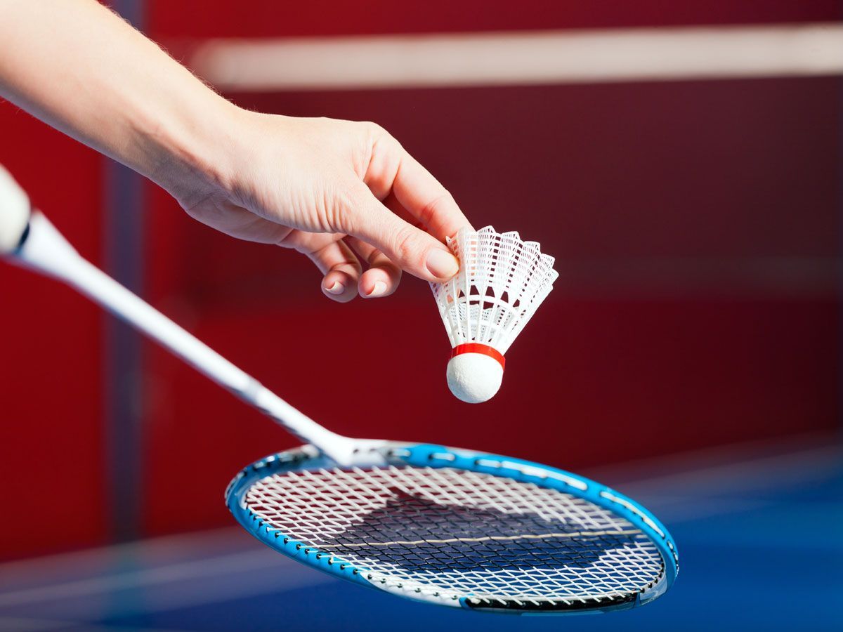 A hand holds a shuttlecock above a blue badminton racket with a red and blue blurred background.