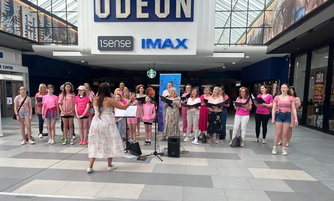 A choir singing in a mall lobby in front of an Odeon cinema, led by a conductor standing with a music stand.