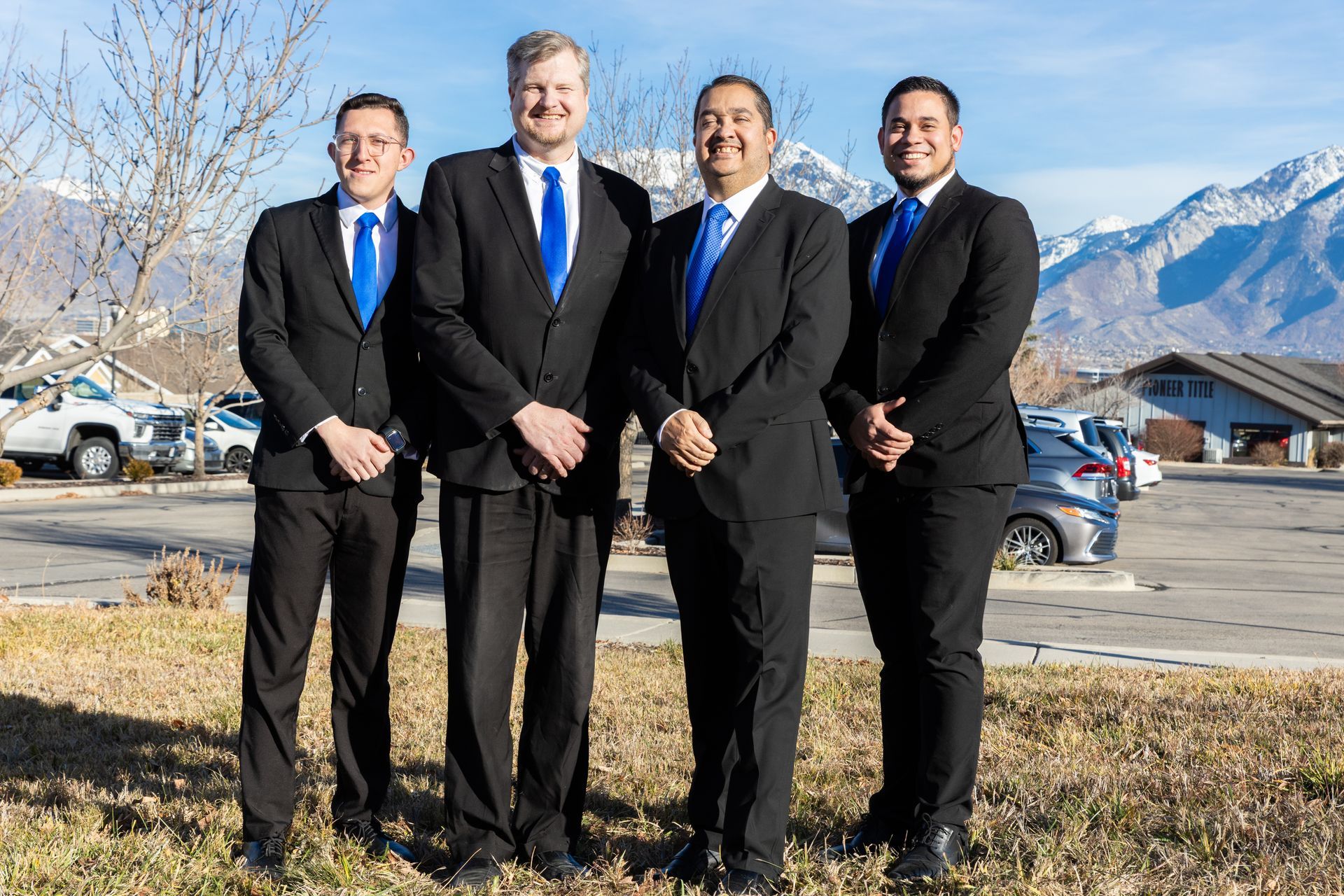 Four men in black suits and blue ties stand outdoors in front of mountains.
