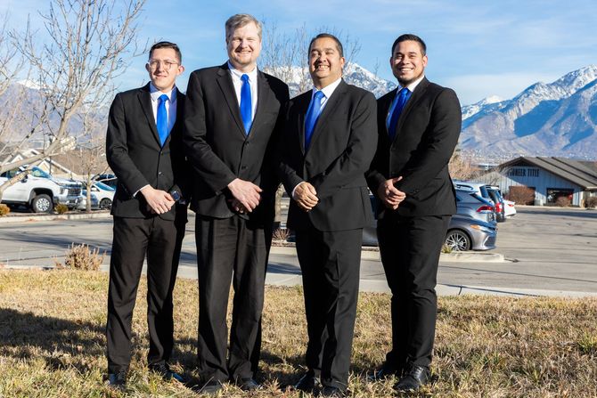 Four men in black suits and blue ties stand outdoors in front of mountains.