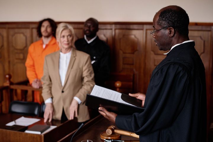 Judge reading from a document in courtroom; defense attorney, defendant, and guard stand by.