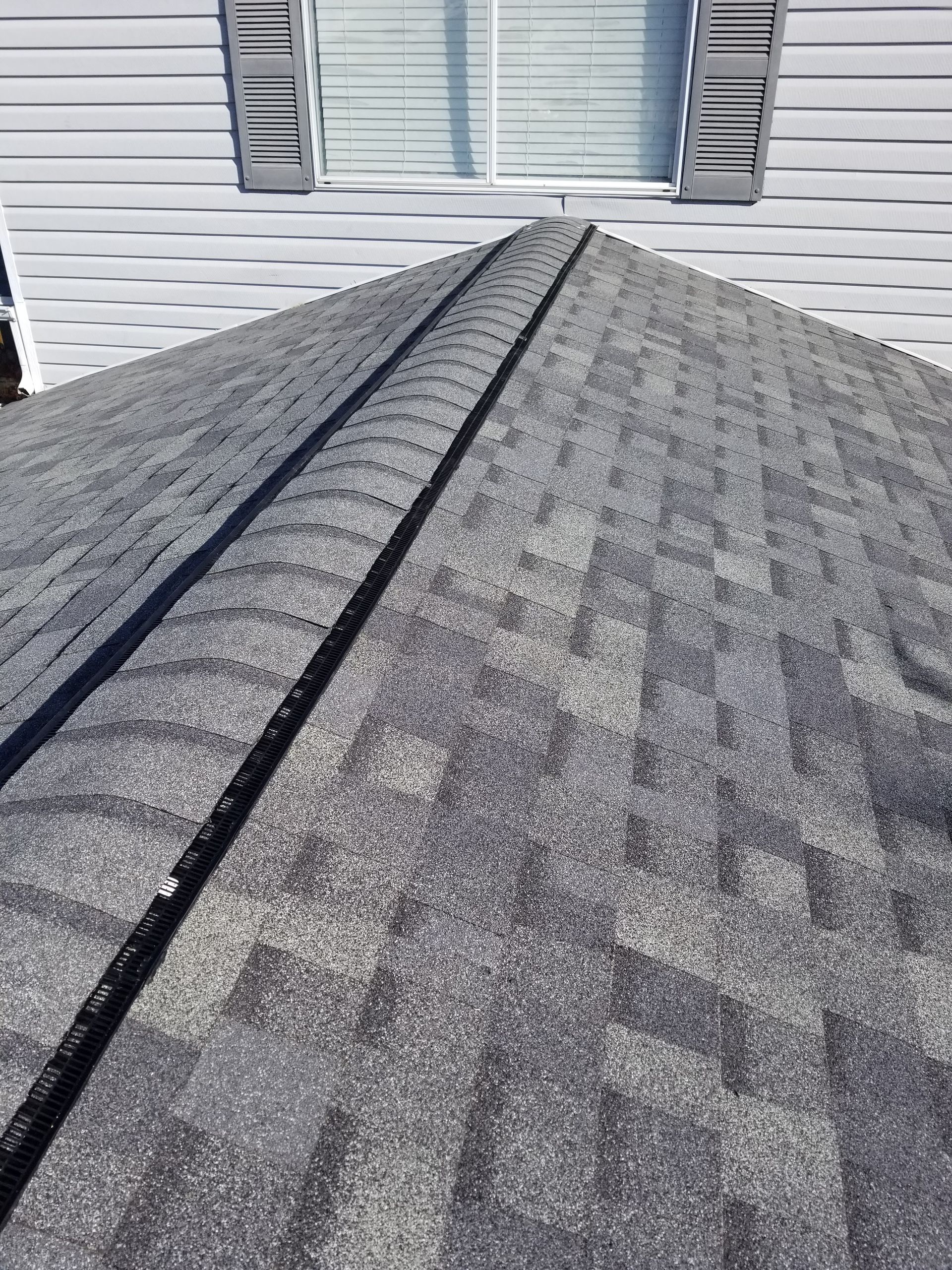 Gray shingled roof with a central seam, viewed from the peak toward a white house wall and windows