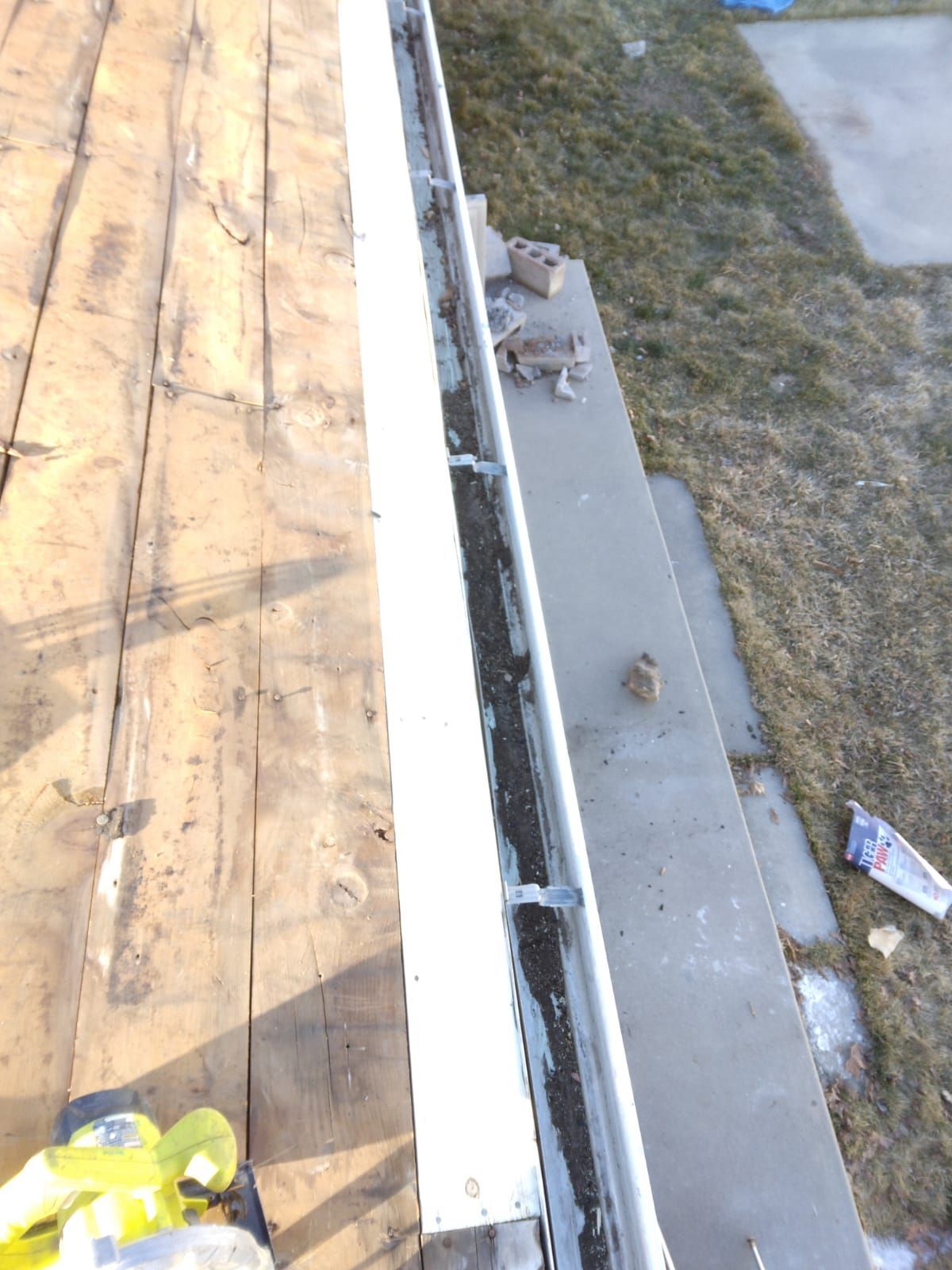Roof edge with exposed white gutter, plywood decking, and debris on the ground below.
