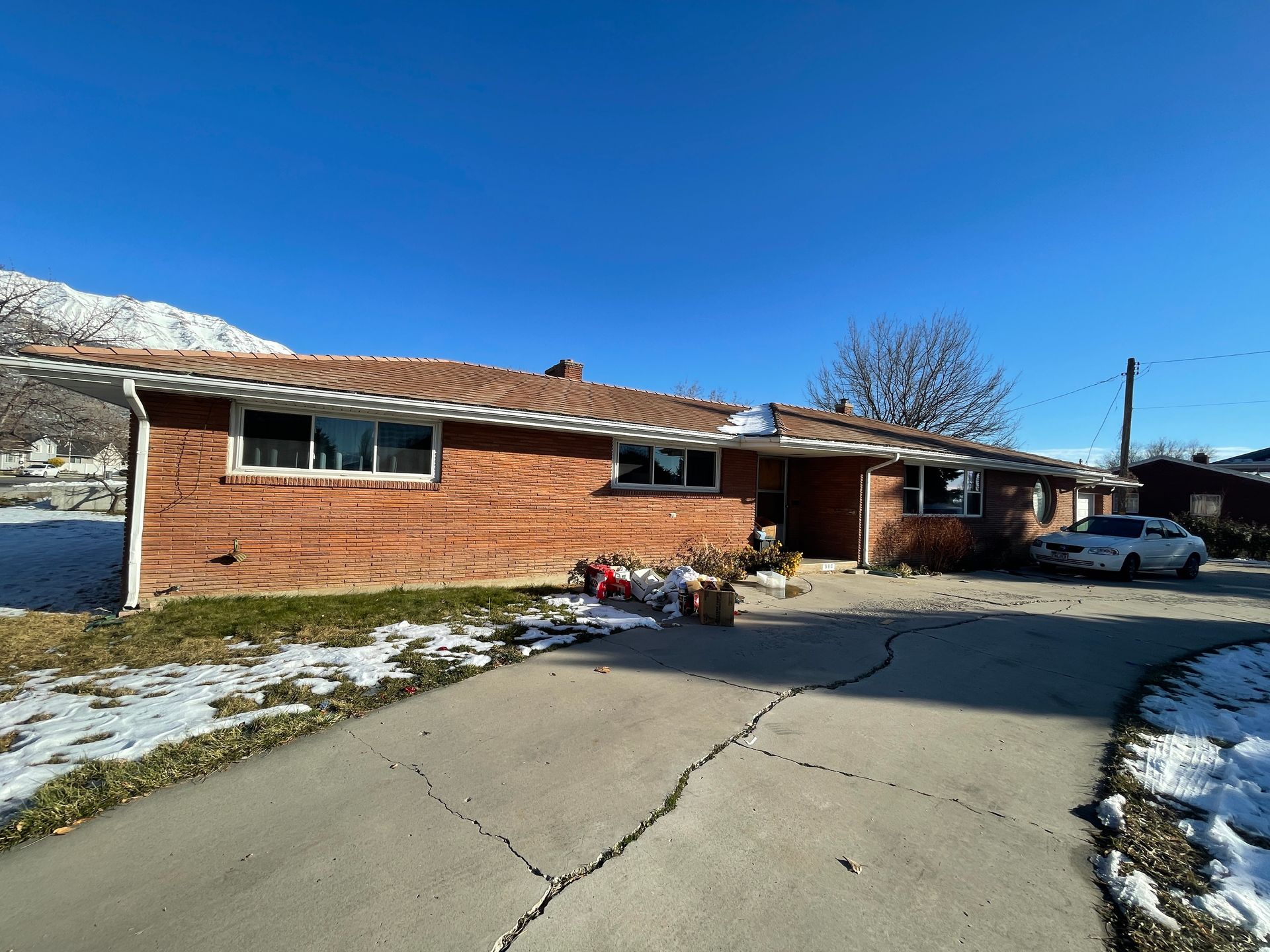Single-story brick house with a curved driveway, patches of snow, and a blue car parked outside.