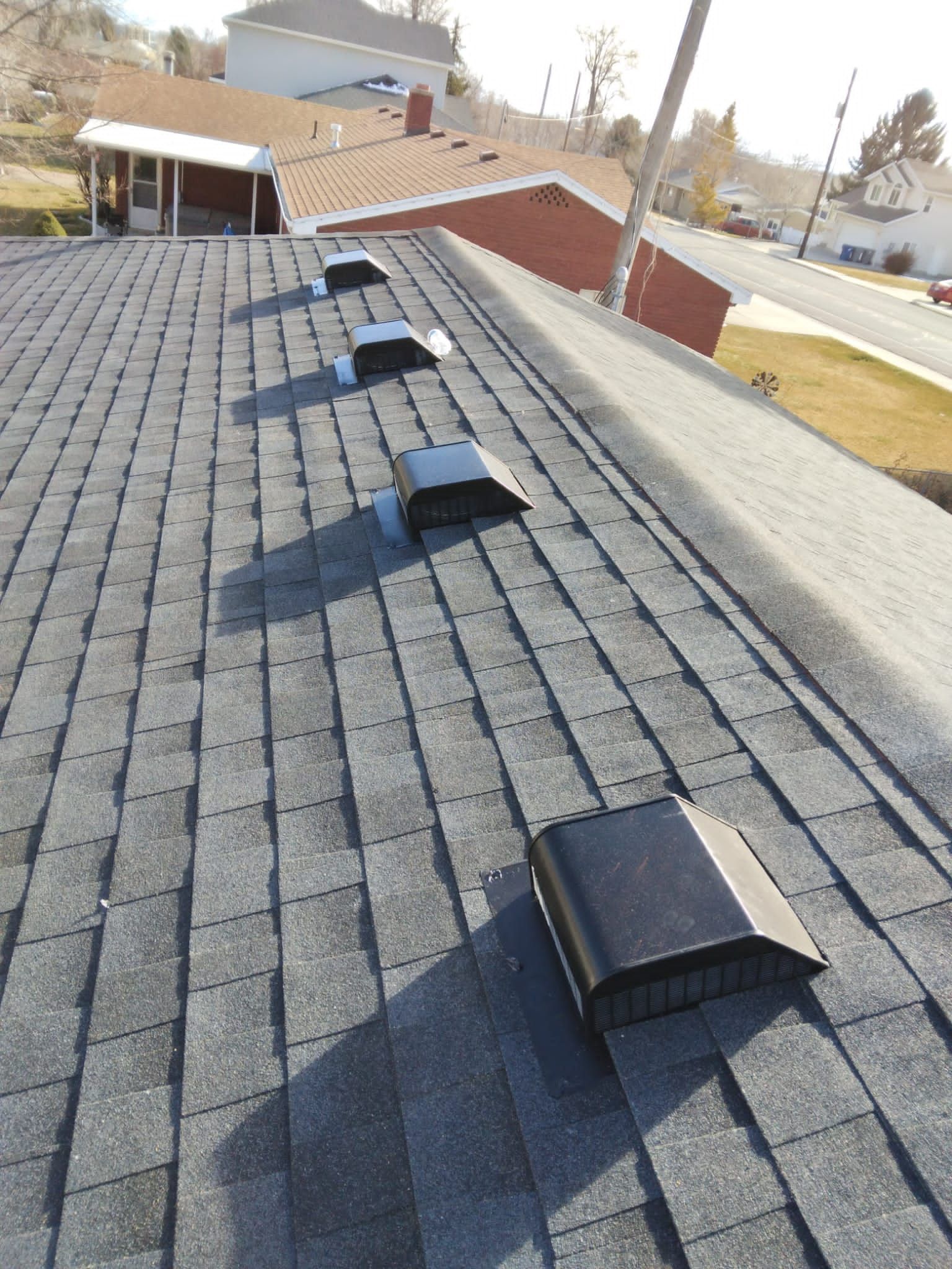 Rooftop with several black roof vents on gray shingles, viewed toward nearby houses and trees.
