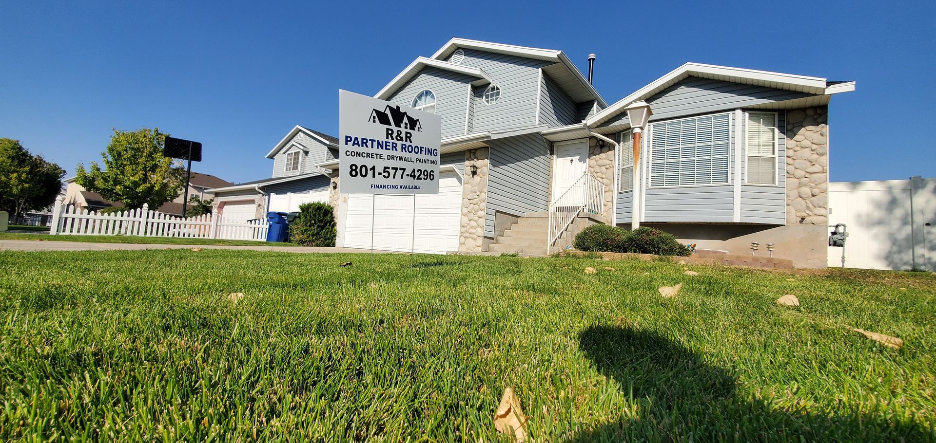 Suburban house with a white fence and sign, viewed from a grassy lawn on a sunny day
