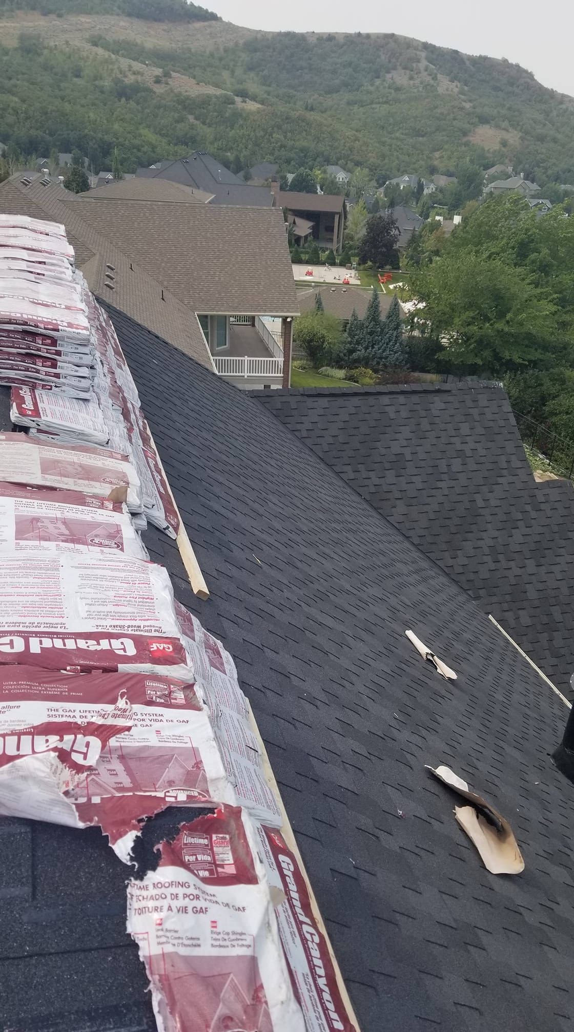 Roof under repair with dark shingles and exposed underlayment, overlooking green hills and distant houses
