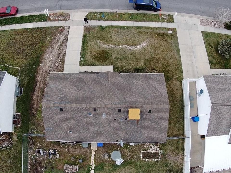 Aerial view of a house with a gray roof, driveway, and grassy yard beside neighboring homes and a street.