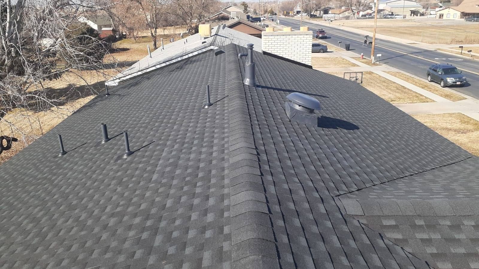 Aerial view of a dark shingle roof with vents and nearby road, houses, and trees in the background.