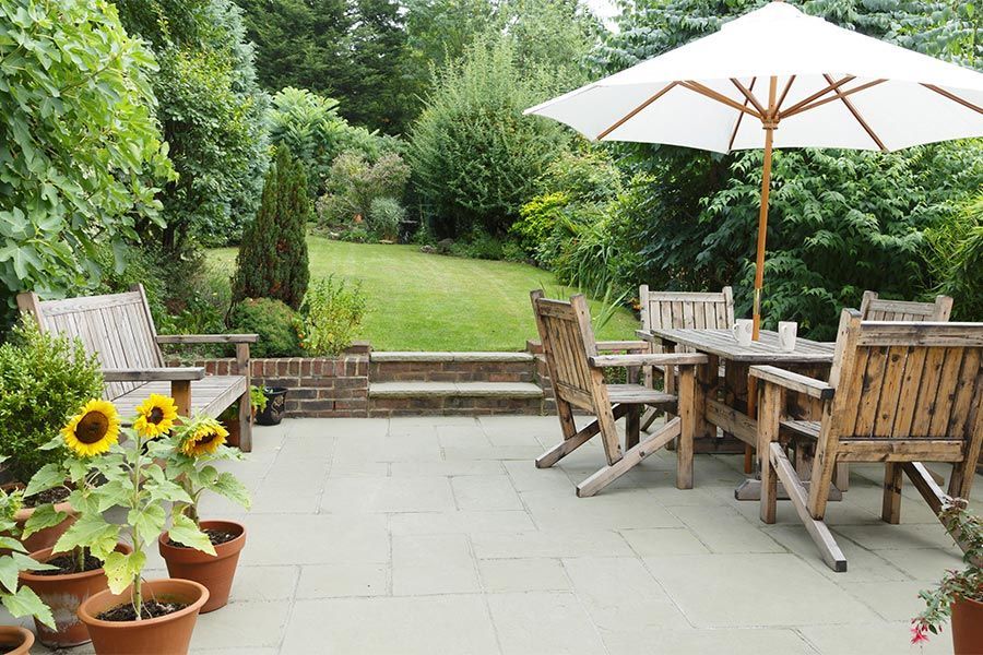 Patio with wooden table and chairs under a white umbrella, potted sunflowers, and a green garden beyond