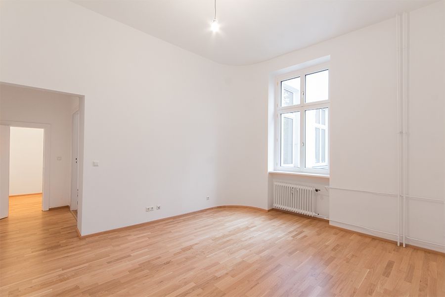 Empty white room with hardwood floors, large window, and radiator by the wall