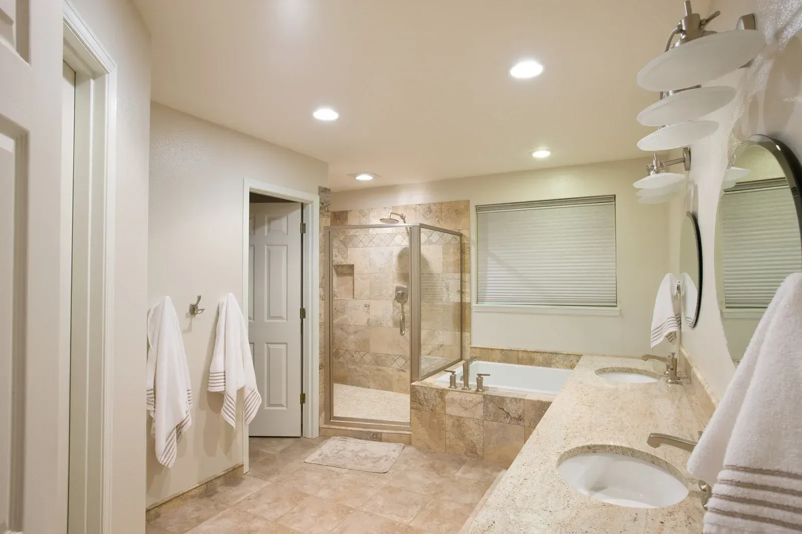 Luxury beige bathroom with double sinks, glass shower, soaking tub, and large window blinds