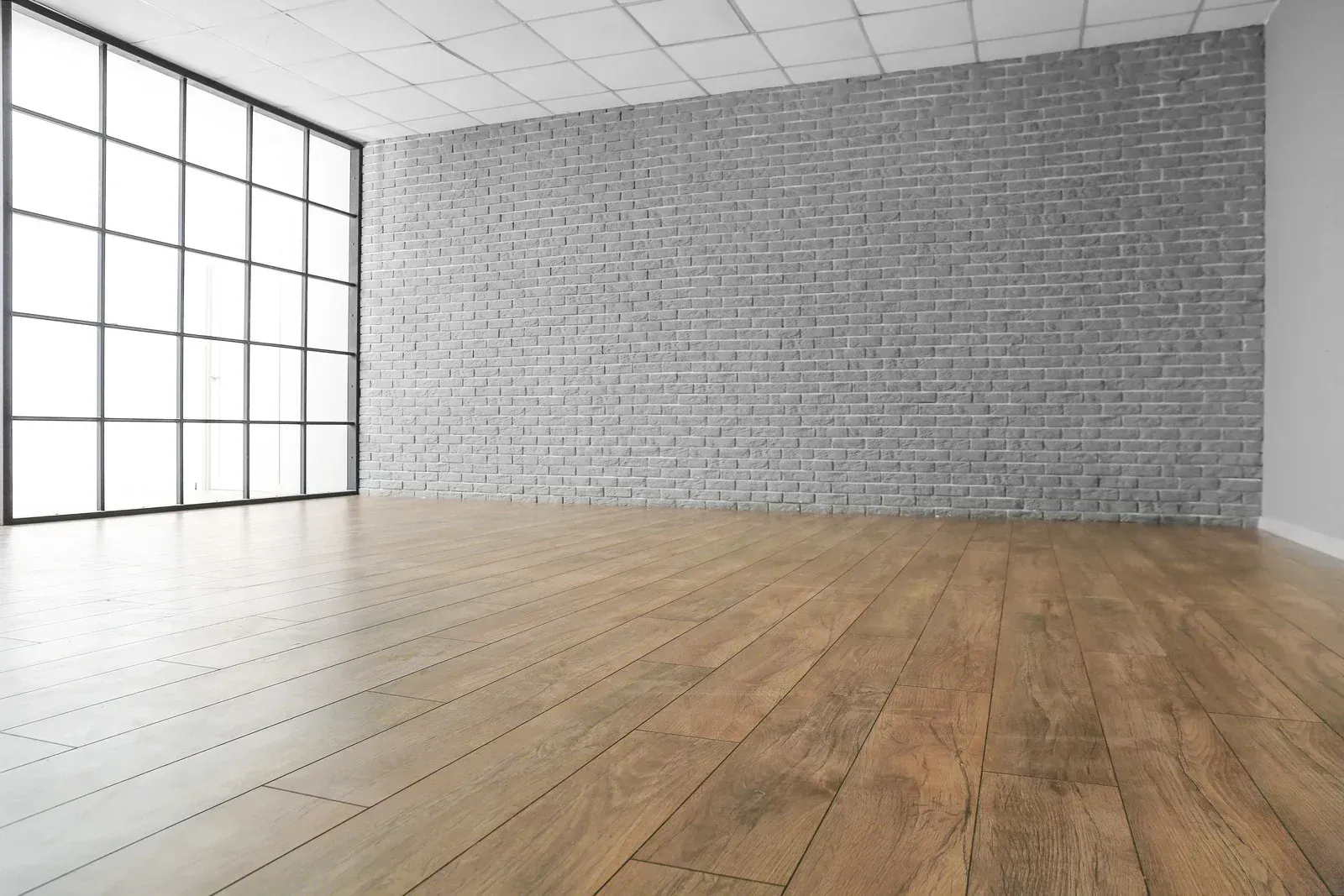 Empty modern loft with large grid windows, white ceiling, exposed brick wall, and light wood floor.