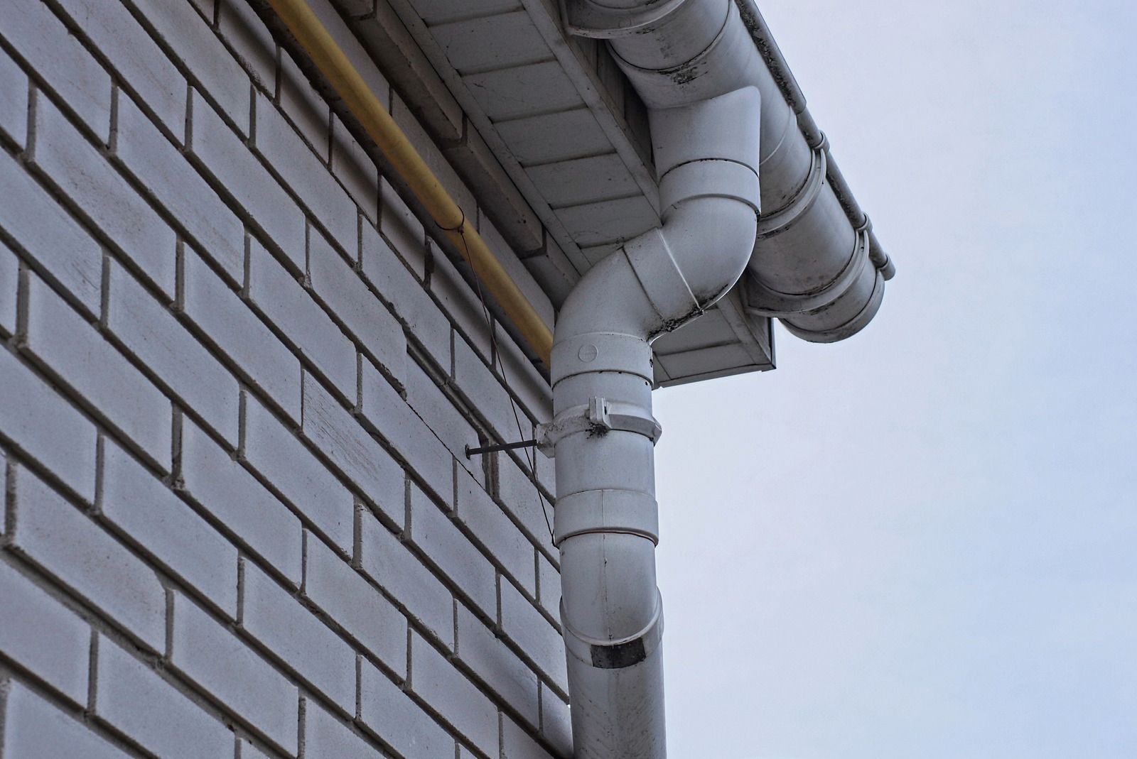 White drainpipe with elbow joints on a brick house wall against a cloudy sky