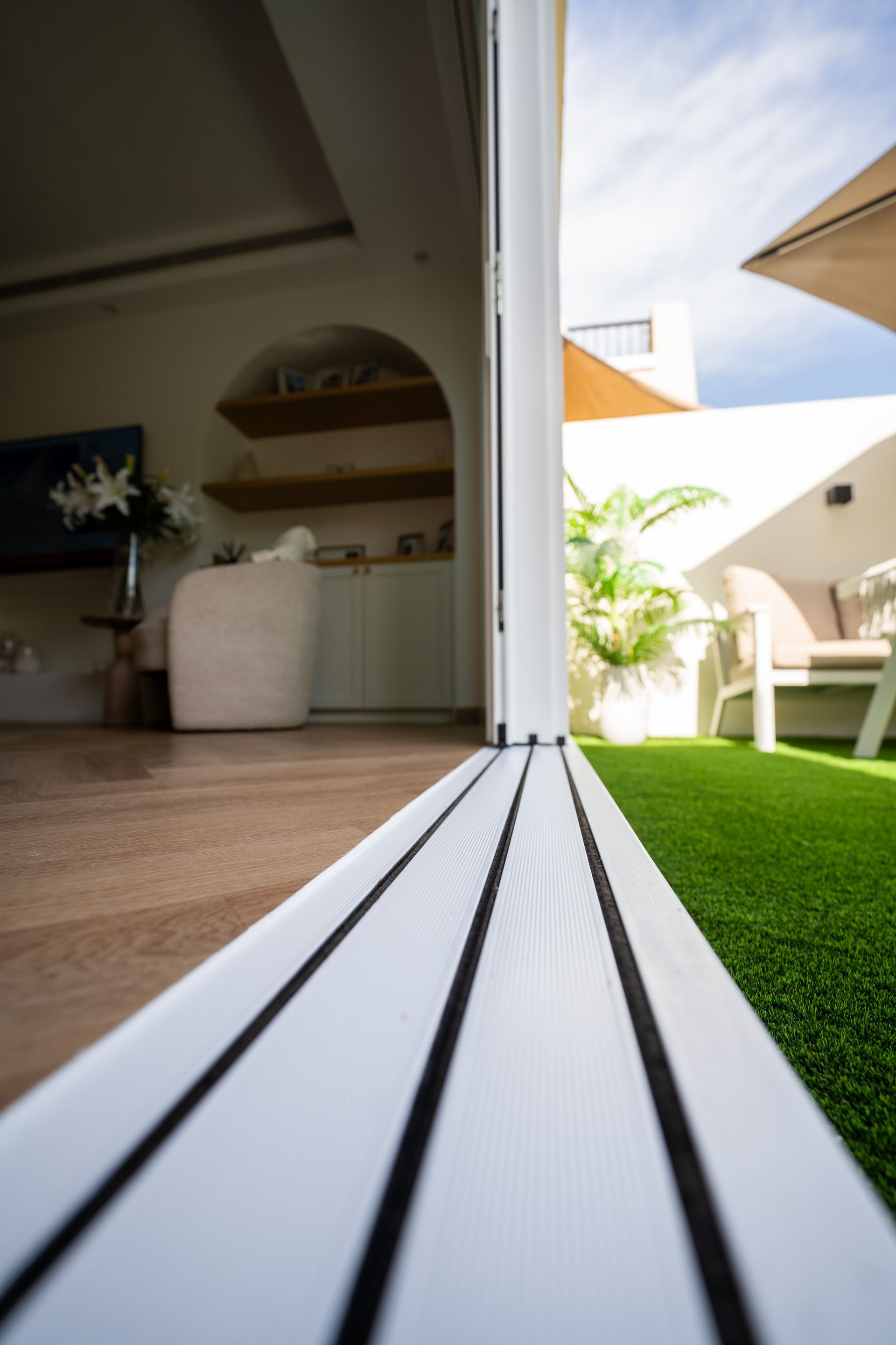 Indoor-outdoor view through open sliding doors to a sunny patio with white chairs, potted plants, and green turf.