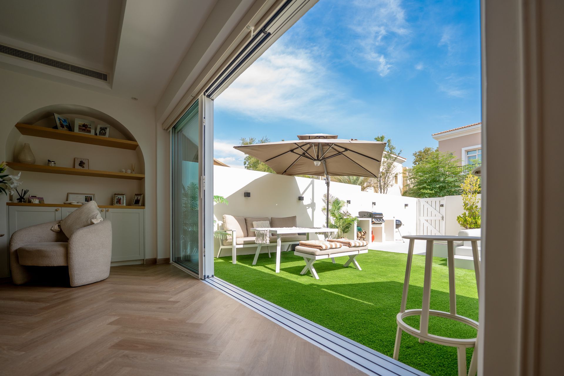 Living room opening to a patio with green turf, outdoor table, chairs, and umbrella under a blue sky