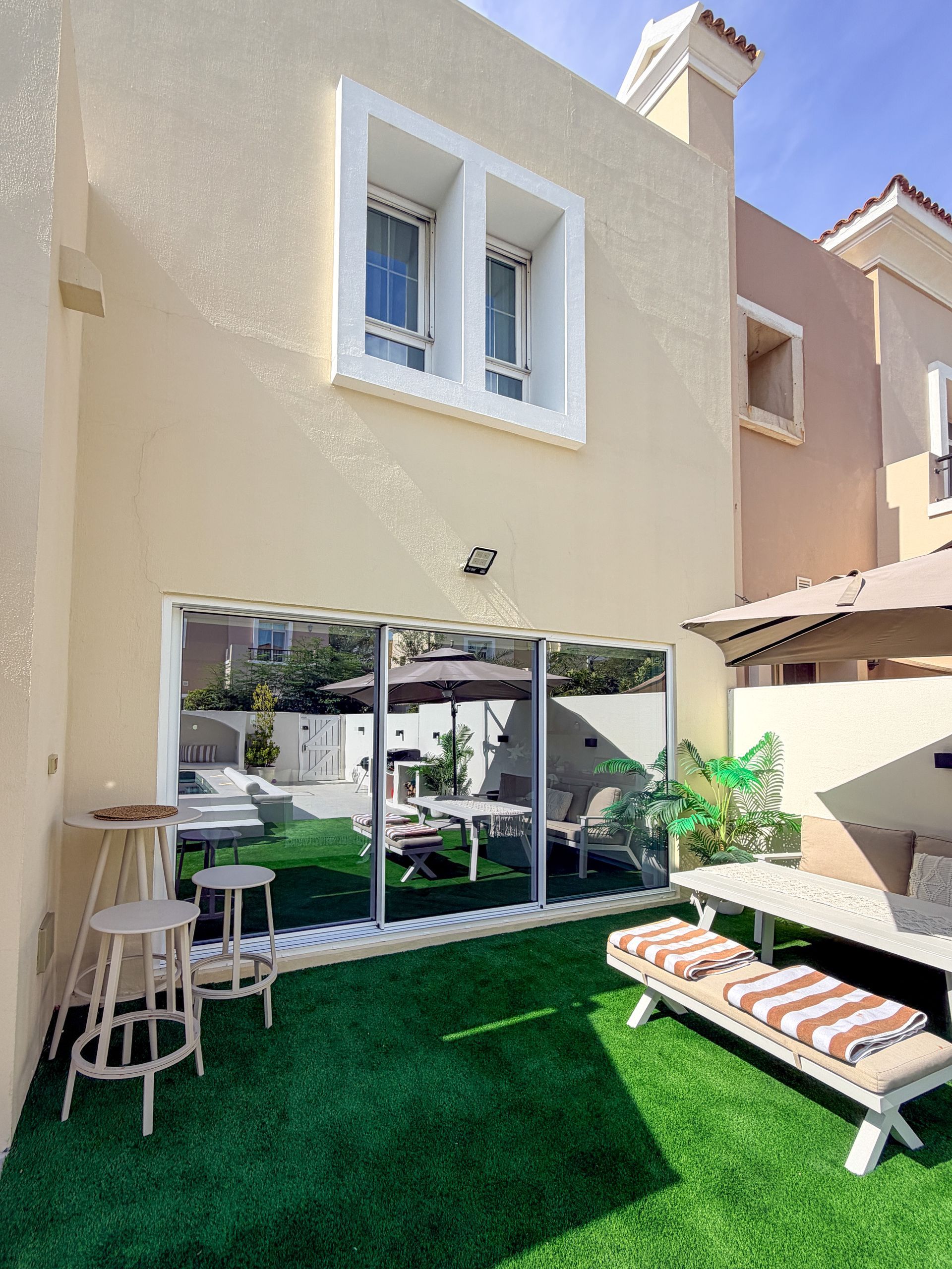 Backyard patio with glass doors, green turf, lounge chair, and small table by a beige house