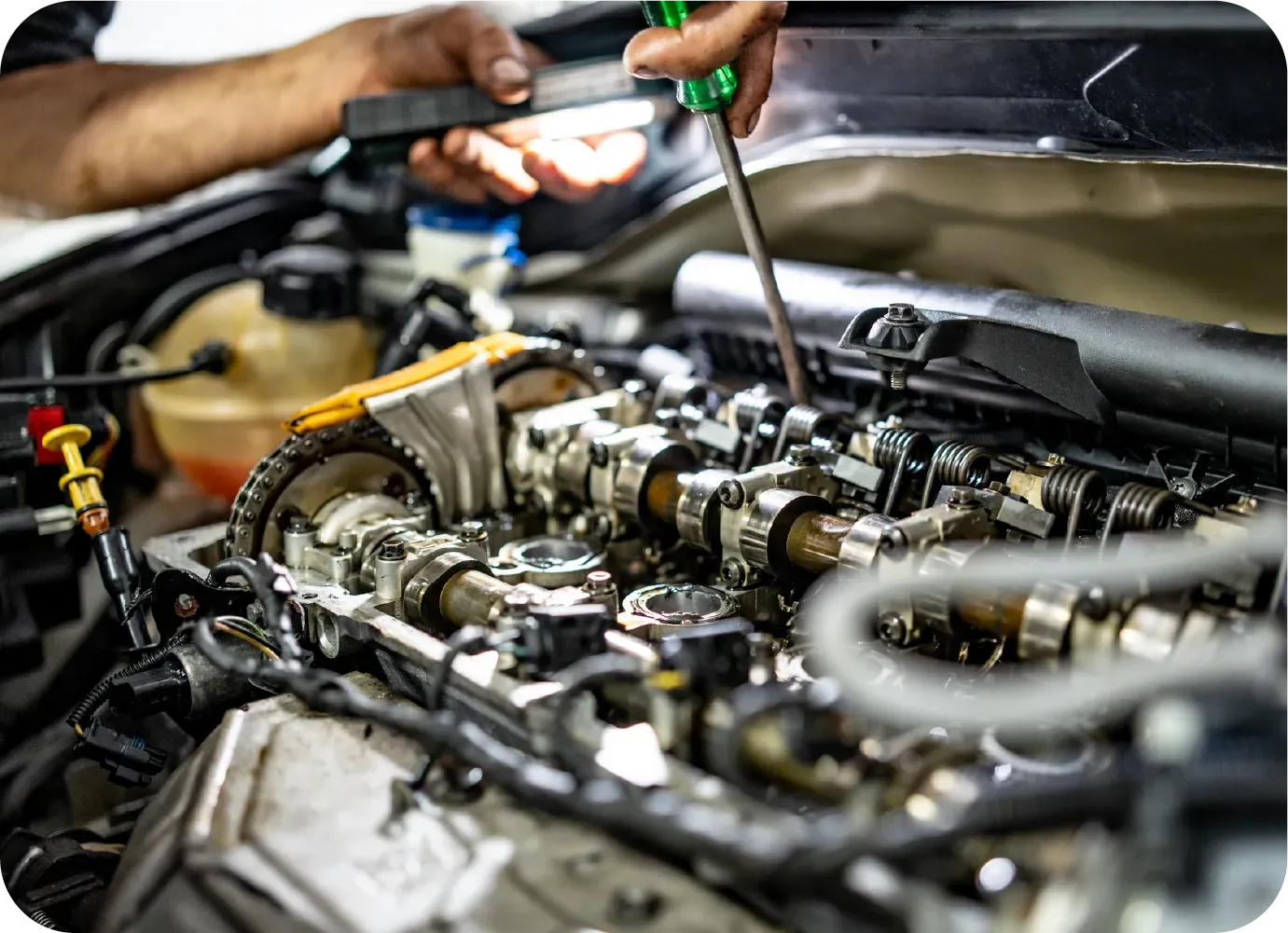 A mechanic uses a screwdriver and a light to inspect the internal camshaft components of an open car engine.