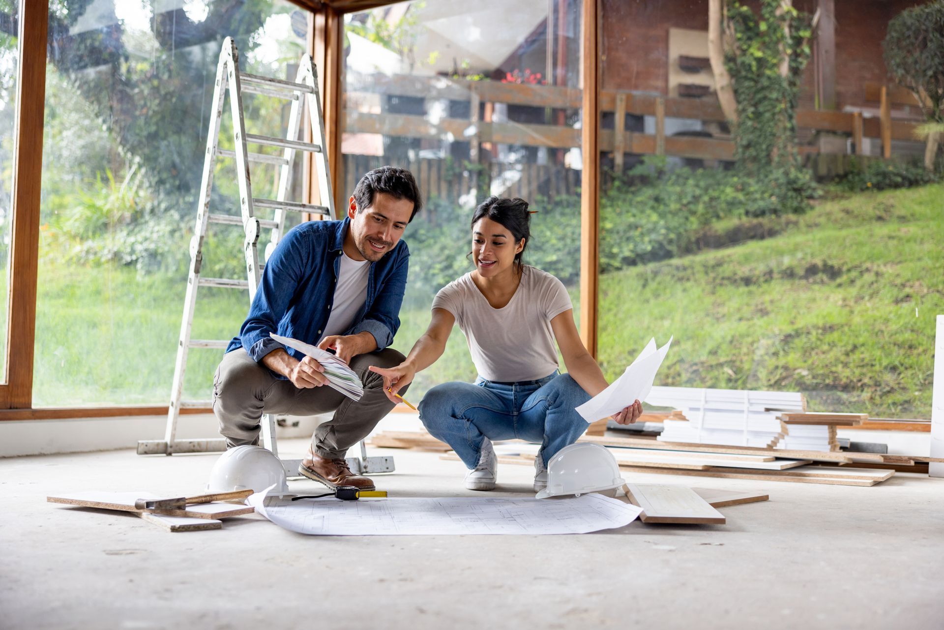 Couple crouches, looking at blueprints in a room under renovation, light-filled with a backyard view.