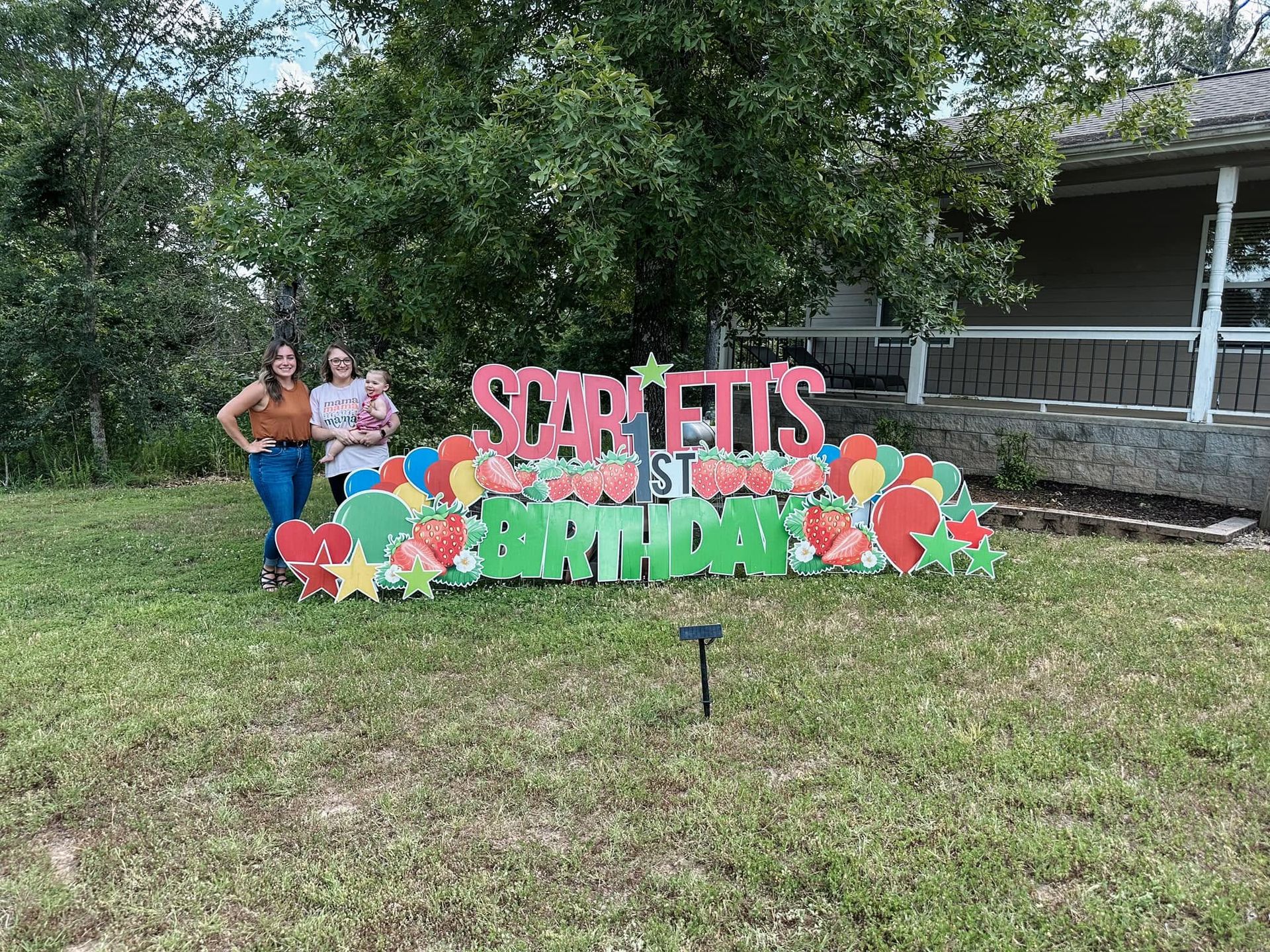 Two people posing by a colorful “SWEETS BIRTHDAY” sign on a grassy lawn with trees and a porch behind them