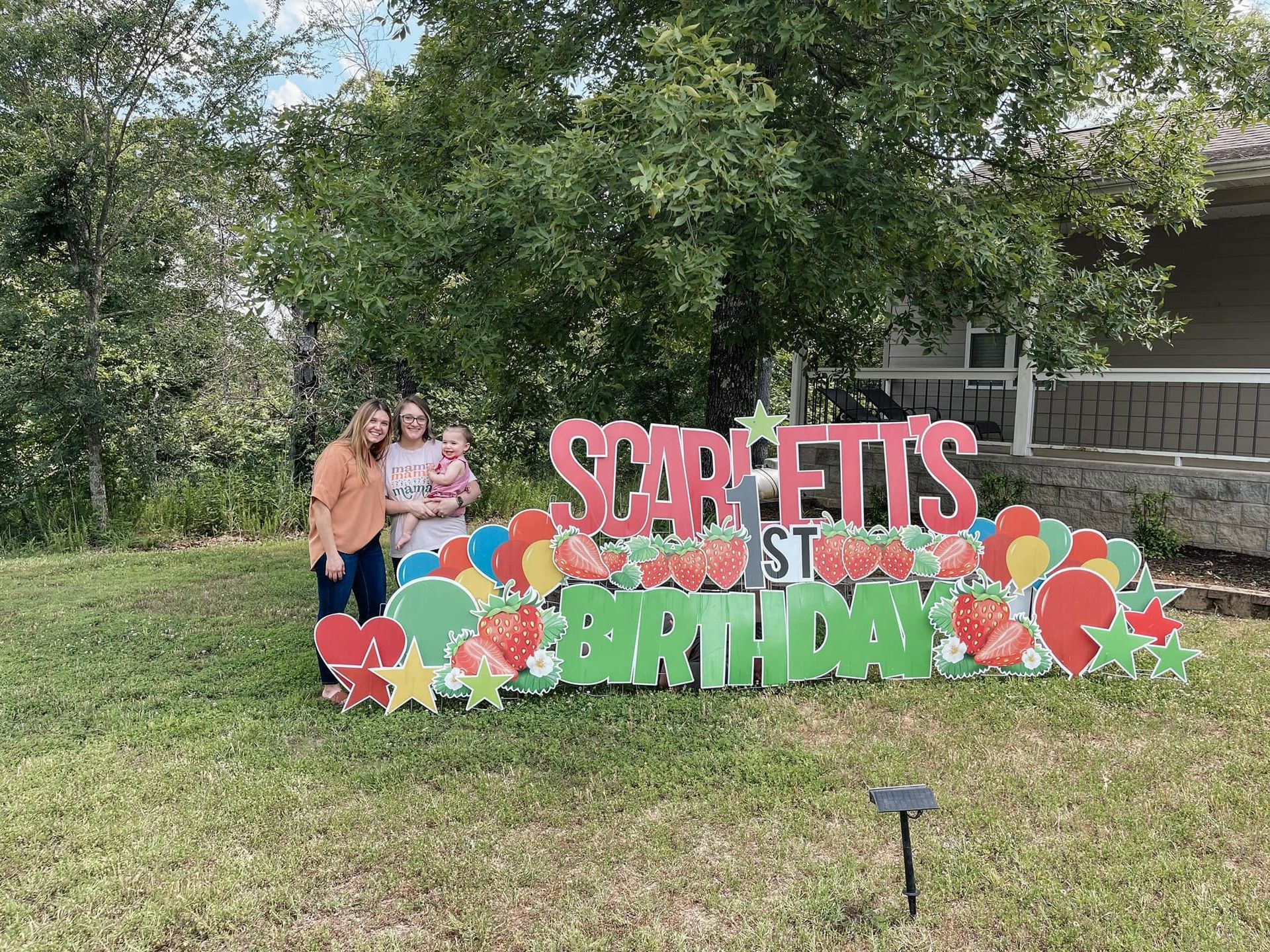 Three people posing behind a colorful “Happy Birthday” yard sign on a grassy lawn.