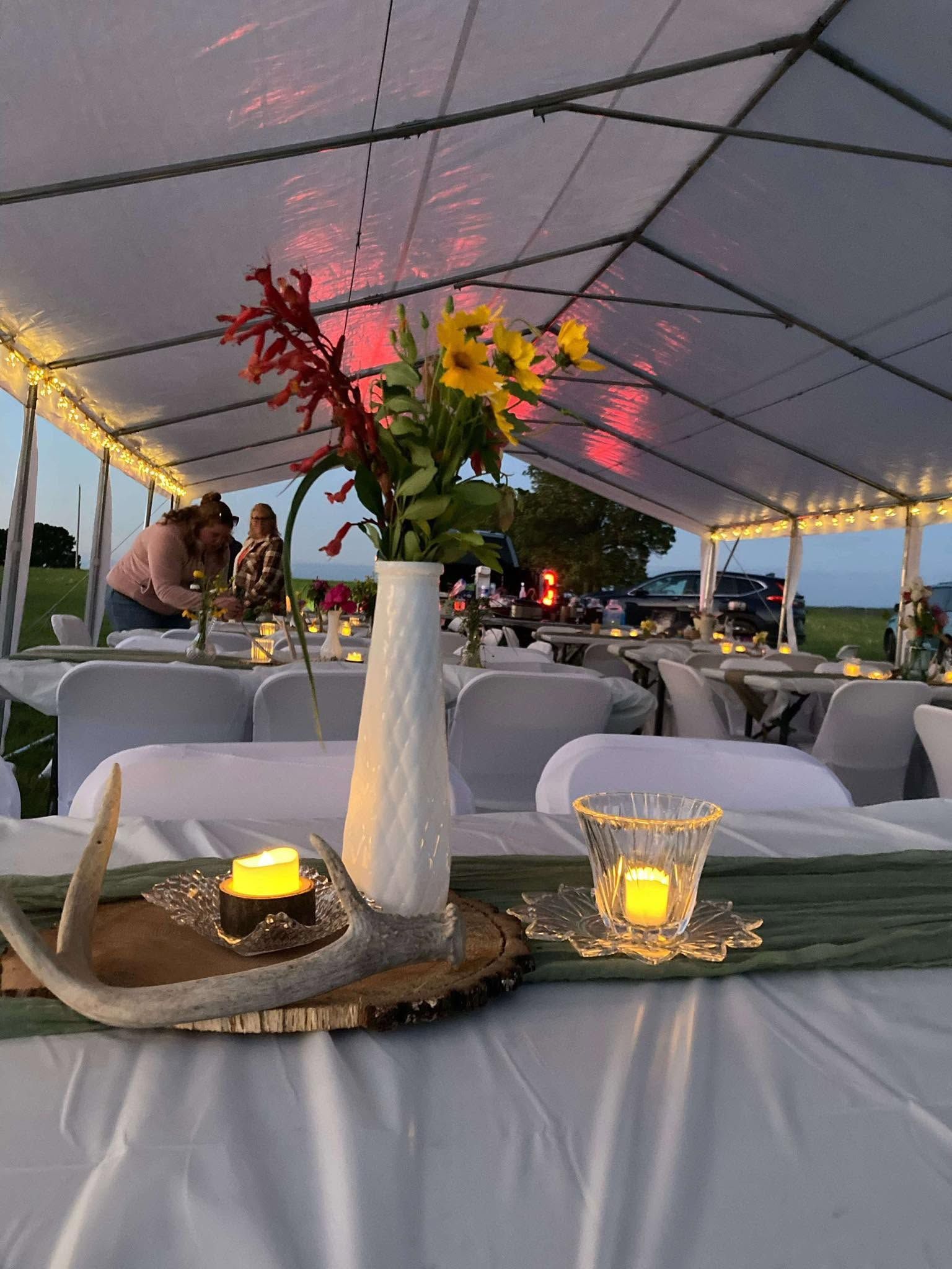 Decorated outdoor event tent with white chairs, candlelit tables, and a floral centerpiece at dusk