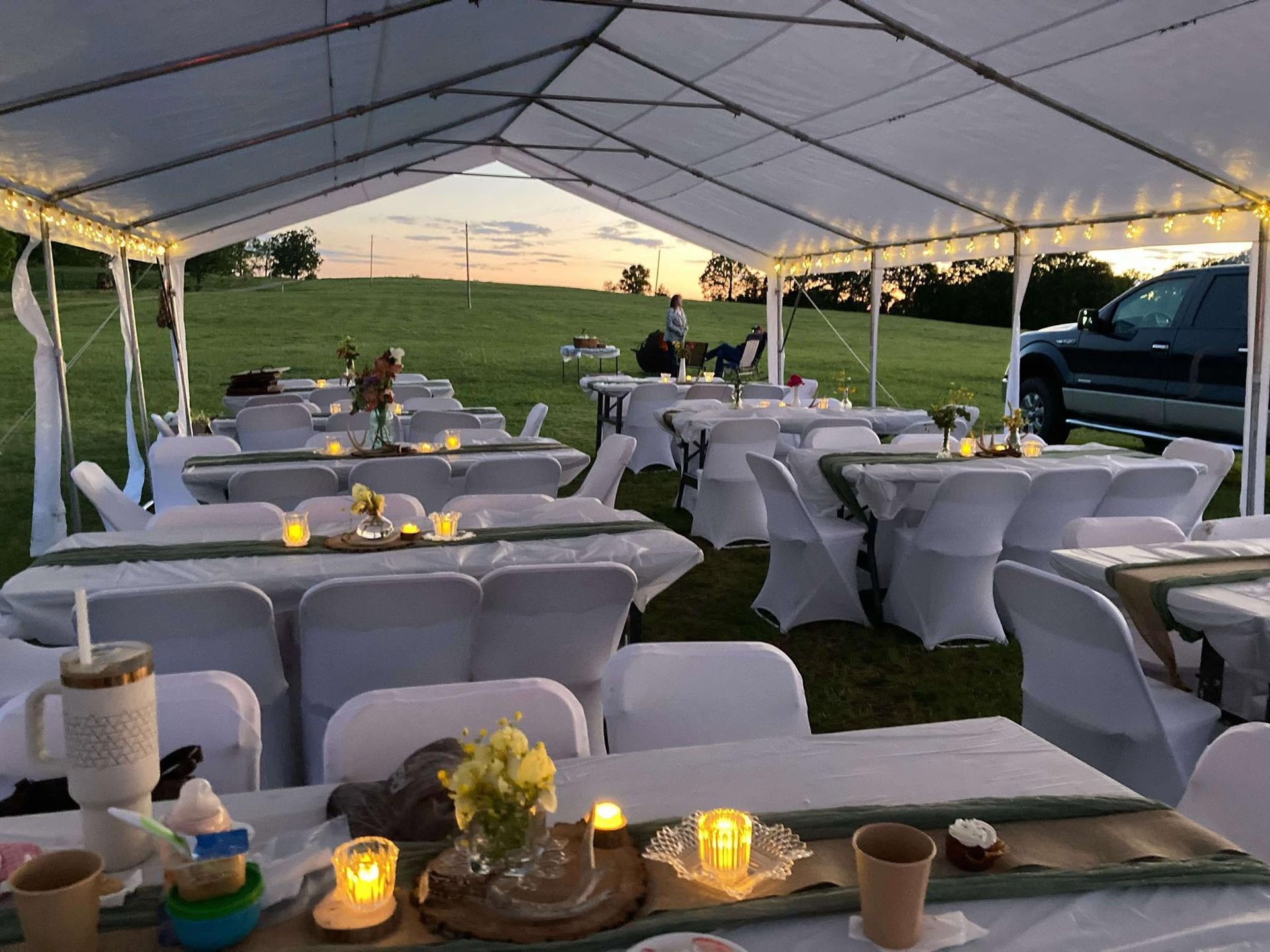 Outdoor event tent with white tables and chairs, lit candles, and a grassy field at sunset