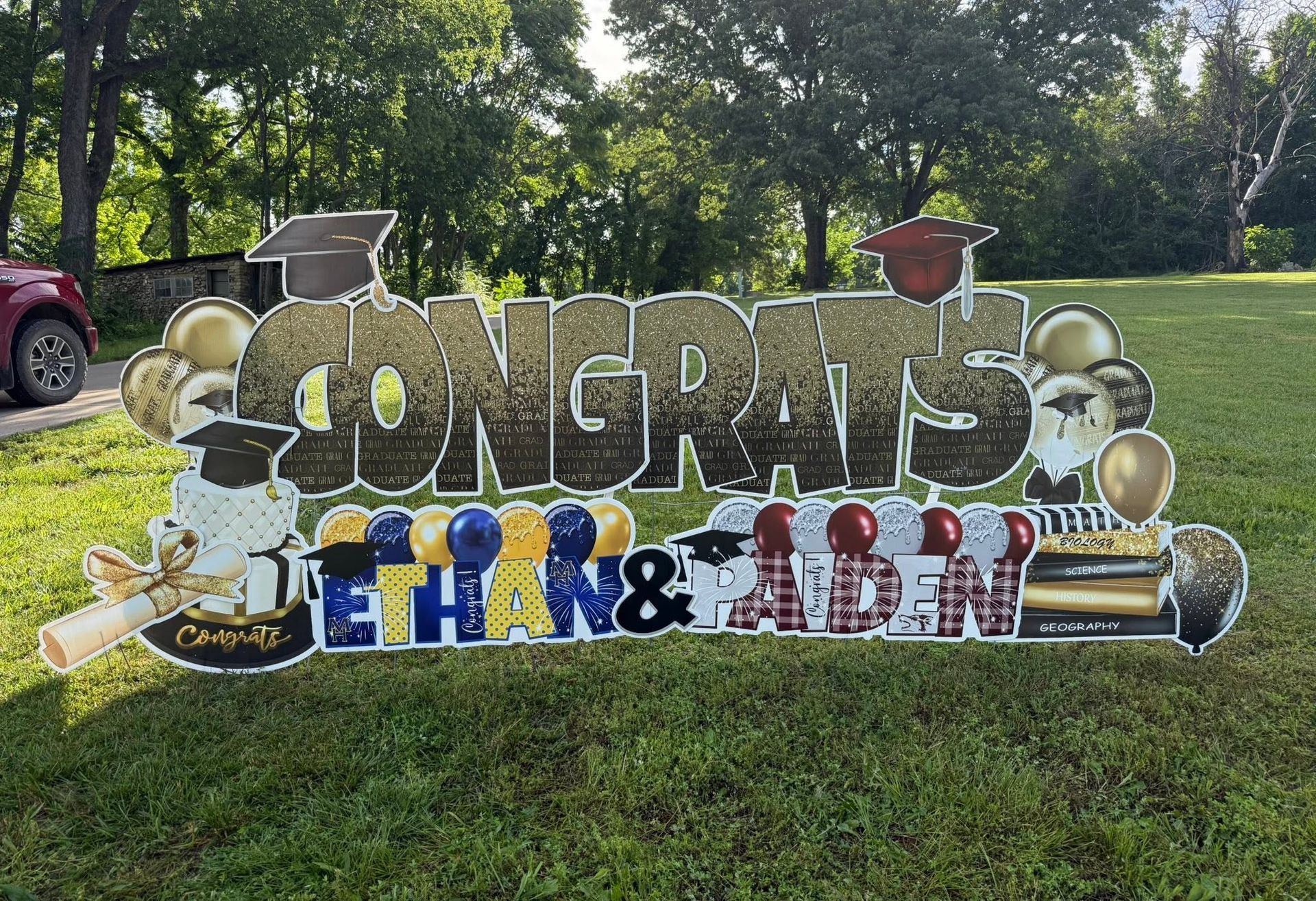 Outdoor congratulatory yard sign with large gold “CONGRATS” letters and blue-red names on a grassy lawn.