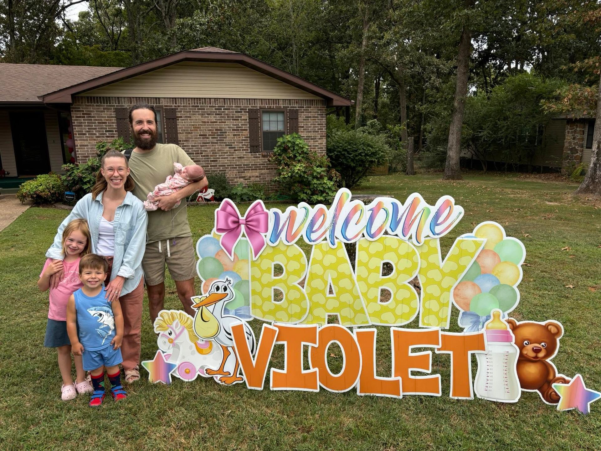 Family posing with pastel “Welcome Baby Violet” sign in front of a house and yard