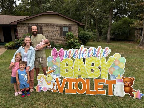 Family posing beside “Welcome Baby Violet” yard sign in front of a house
