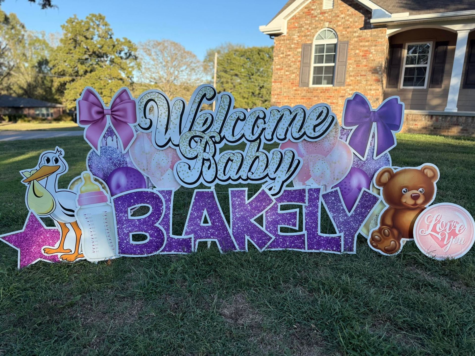 “Welcome Home” purple glitter lawn sign with butterflies and a teddy bear in a front yard