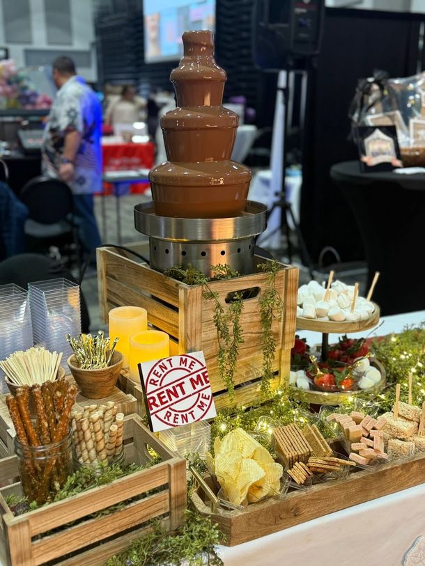 Chocolate fountain dessert display with fruit, marshmallows, and decorative wooden crates at an event booth