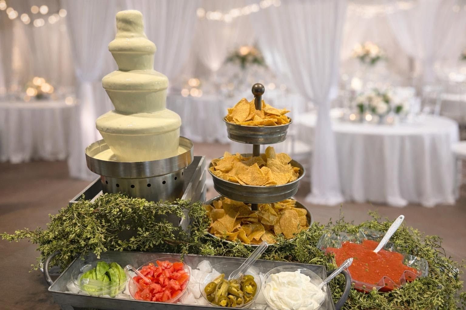 White tiered cake and assorted appetizers on a decorated buffet table in a softly lit banquet room