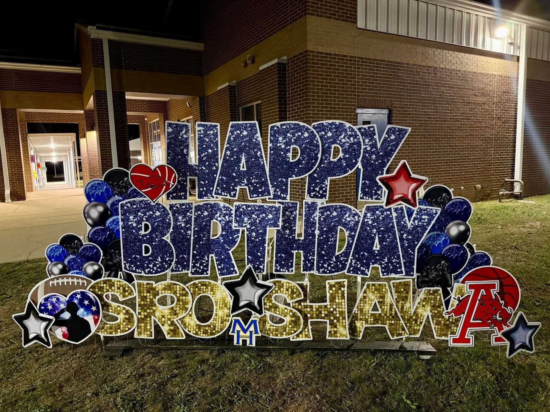 Large blue-and-gold “Happy Birthday” lawn sign with stars outside a brick building at night