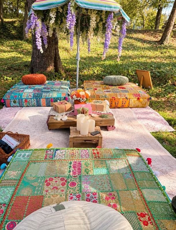 Outdoor picnic setup on patterned rugs with cushions, low tableware, and hanging purple flowers under a canopy.