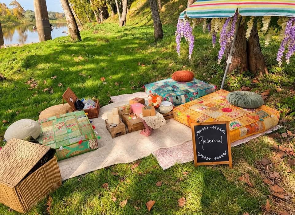 Outdoor picnic setup on grass with colorful cushions, blankets, a chalkboard sign, and hanging flowers by the water