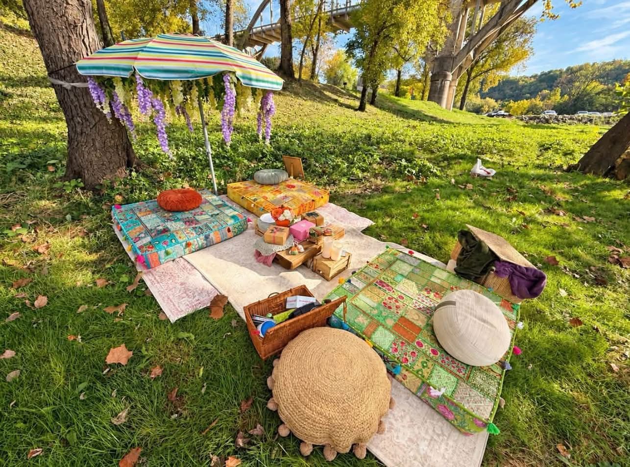 Outdoor picnic setup on patterned rugs with cushions, fruit, bread, and tea under a tree in a sunny garden