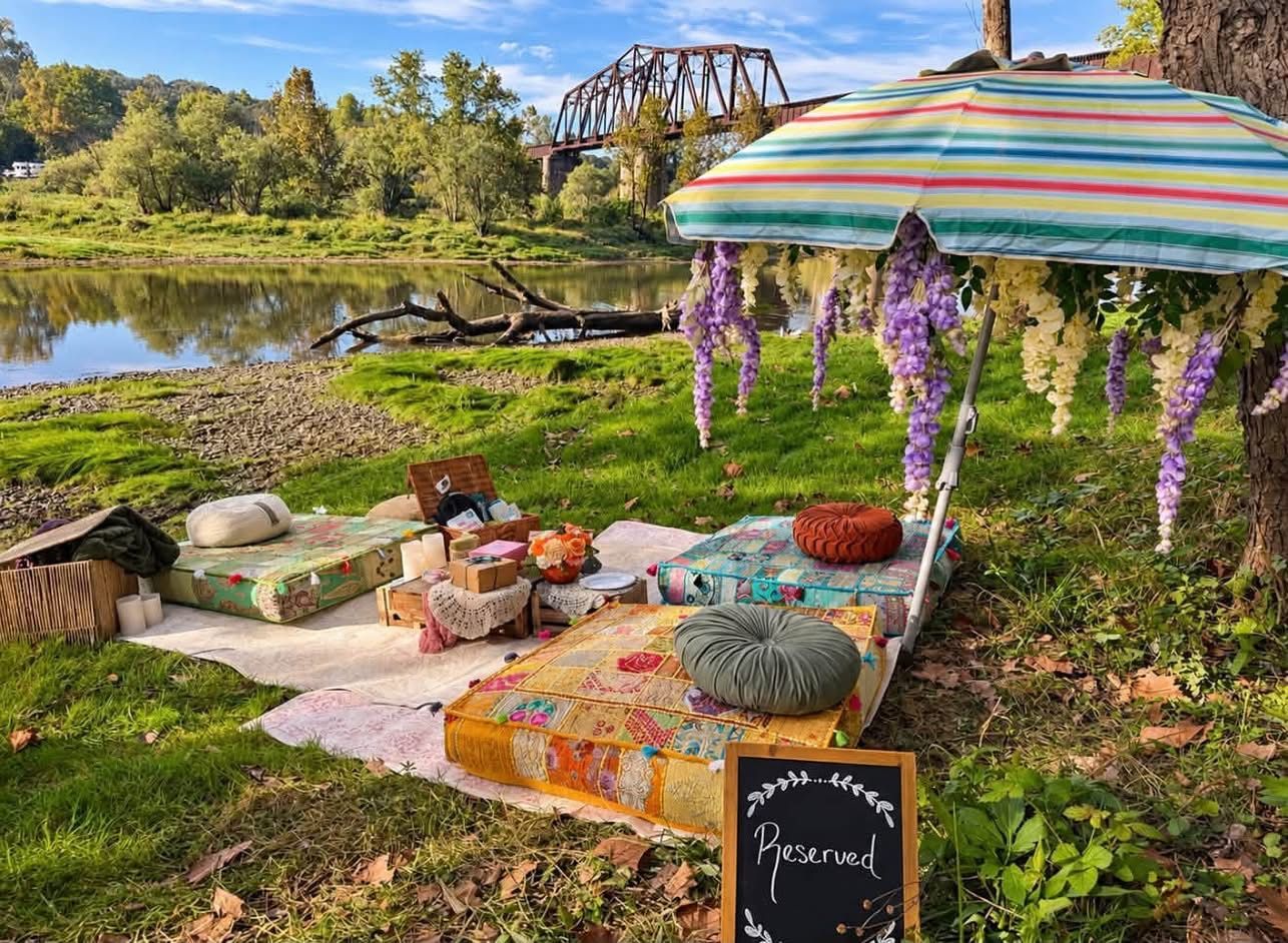 Picnic setup by a river with a striped umbrella, flowers, blankets, and a bridge in the background