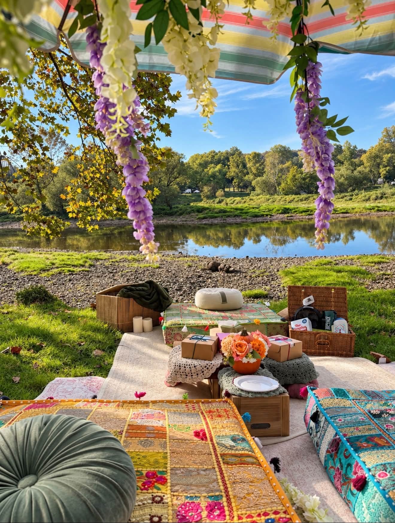 Picnic setup with cushions and flowers under a canopy by a river, framed by colorful floral garlands.