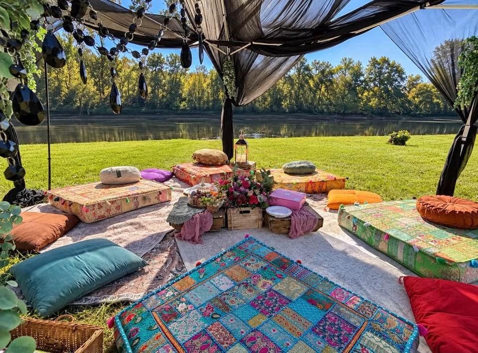Outdoor picnic setup with colorful cushions and low tables under a shaded canopy on a grassy lawn.