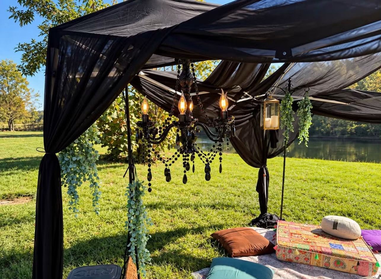 Black-draped outdoor canopy with hanging lights beside a lake, with cushions and a straw hat on a table.