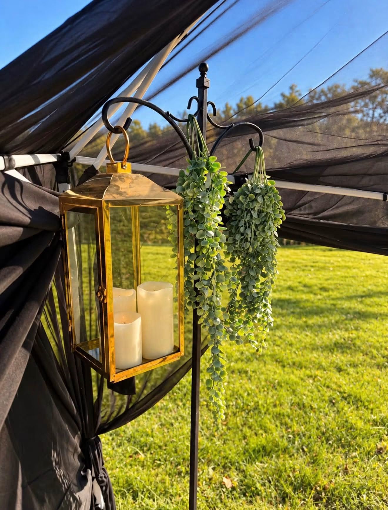 Gold lantern with candles hanging beside a black net tent and green faux greenery outdoors