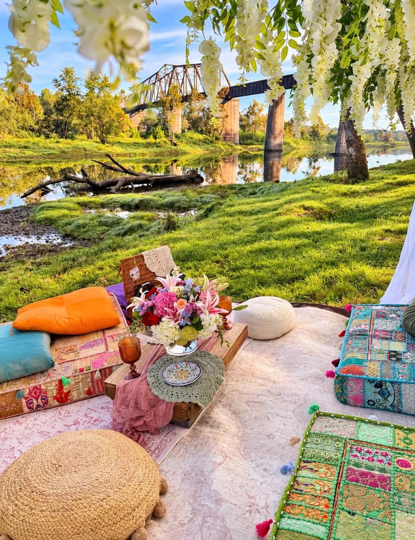 Picnic setup on a blanket by a river, with colorful cushions, flowers, and trees in a sunny park