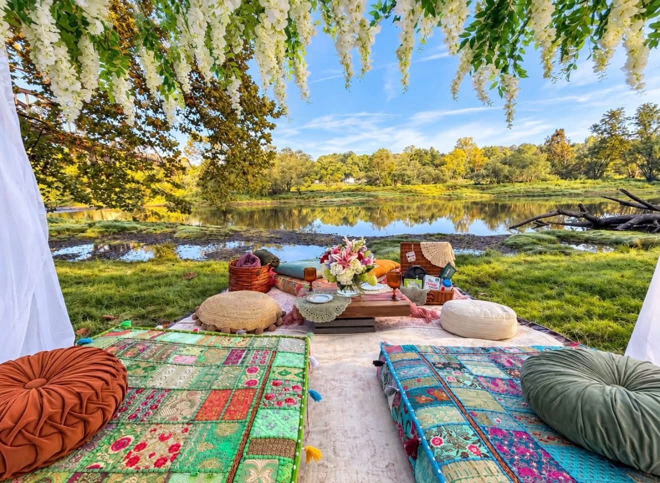 Picnic setup on a blanket by a lake, with cushions, flowers, and trees overhead on a sunny day