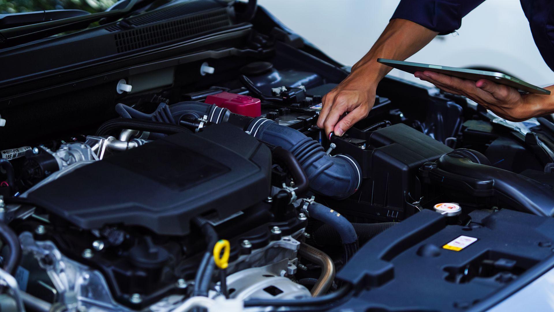 Person inspecting a car engine, holding a tablet.
