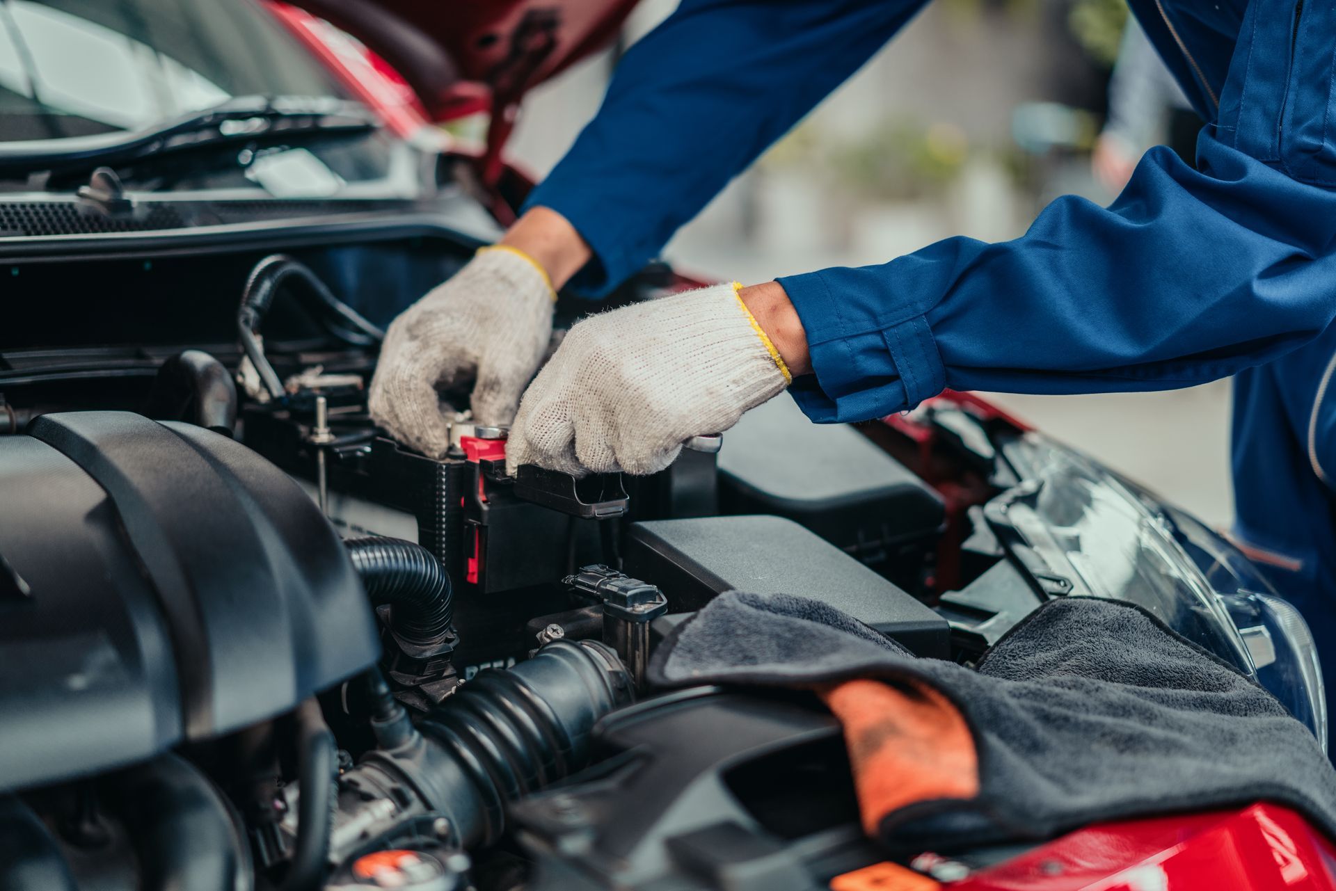 Mechanic in blue coveralls and gloves working on a car engine, connecting wires.
