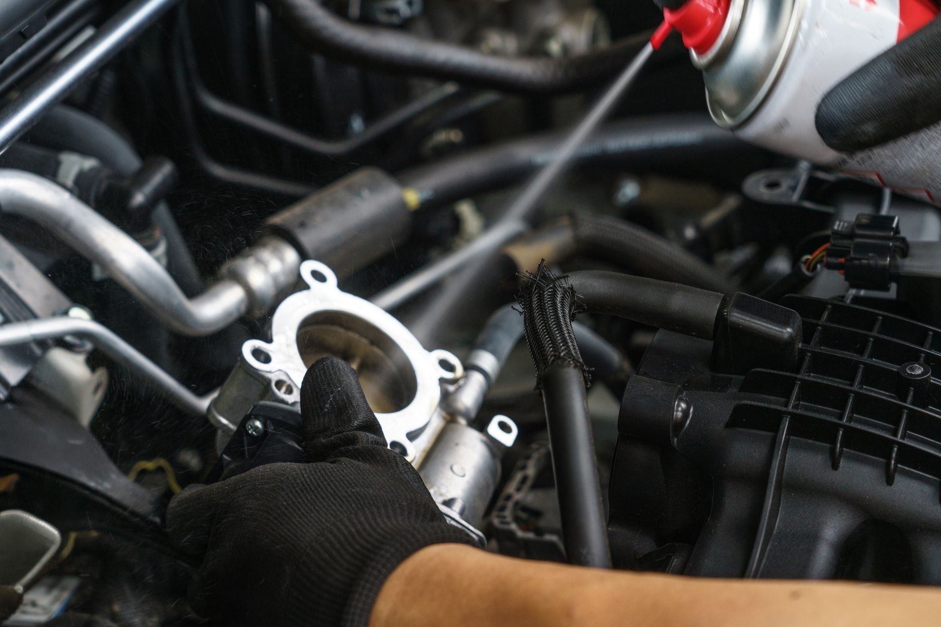 Gloved hand spraying cleaner into a car's throttle body. Engine components visible.