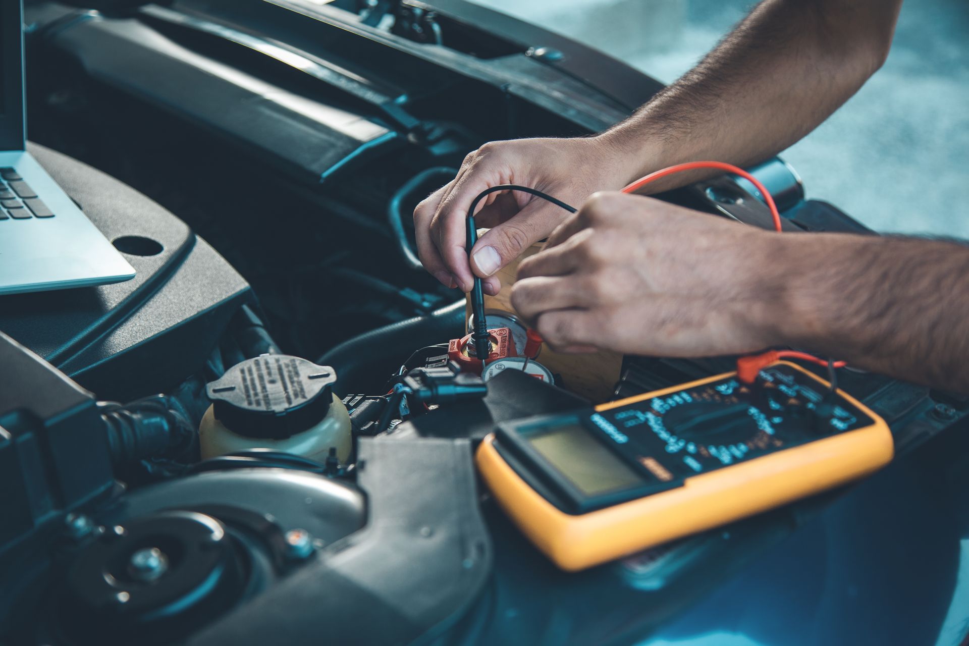 Hands testing a car engine with a multimeter and laptop in an engine bay.
