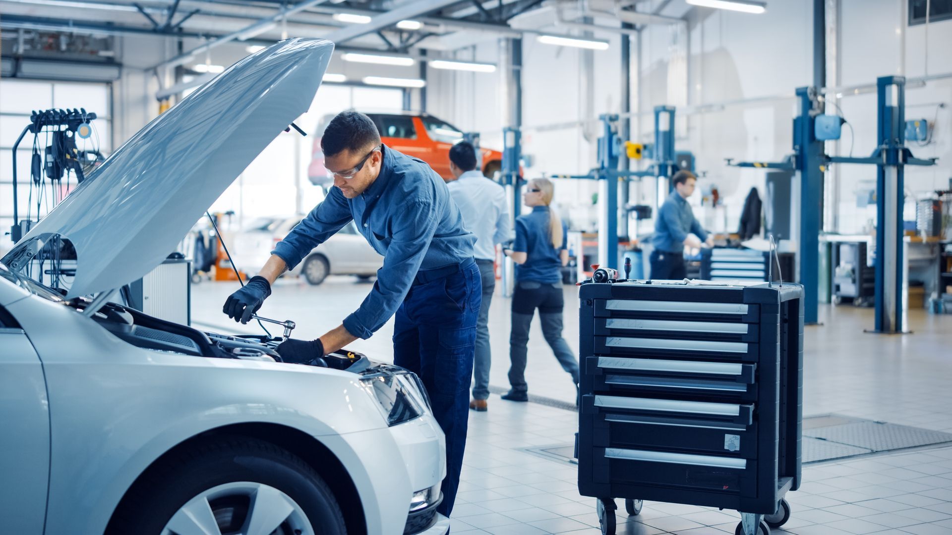 Mechanic working on car engine in a garage with other people and equipment.